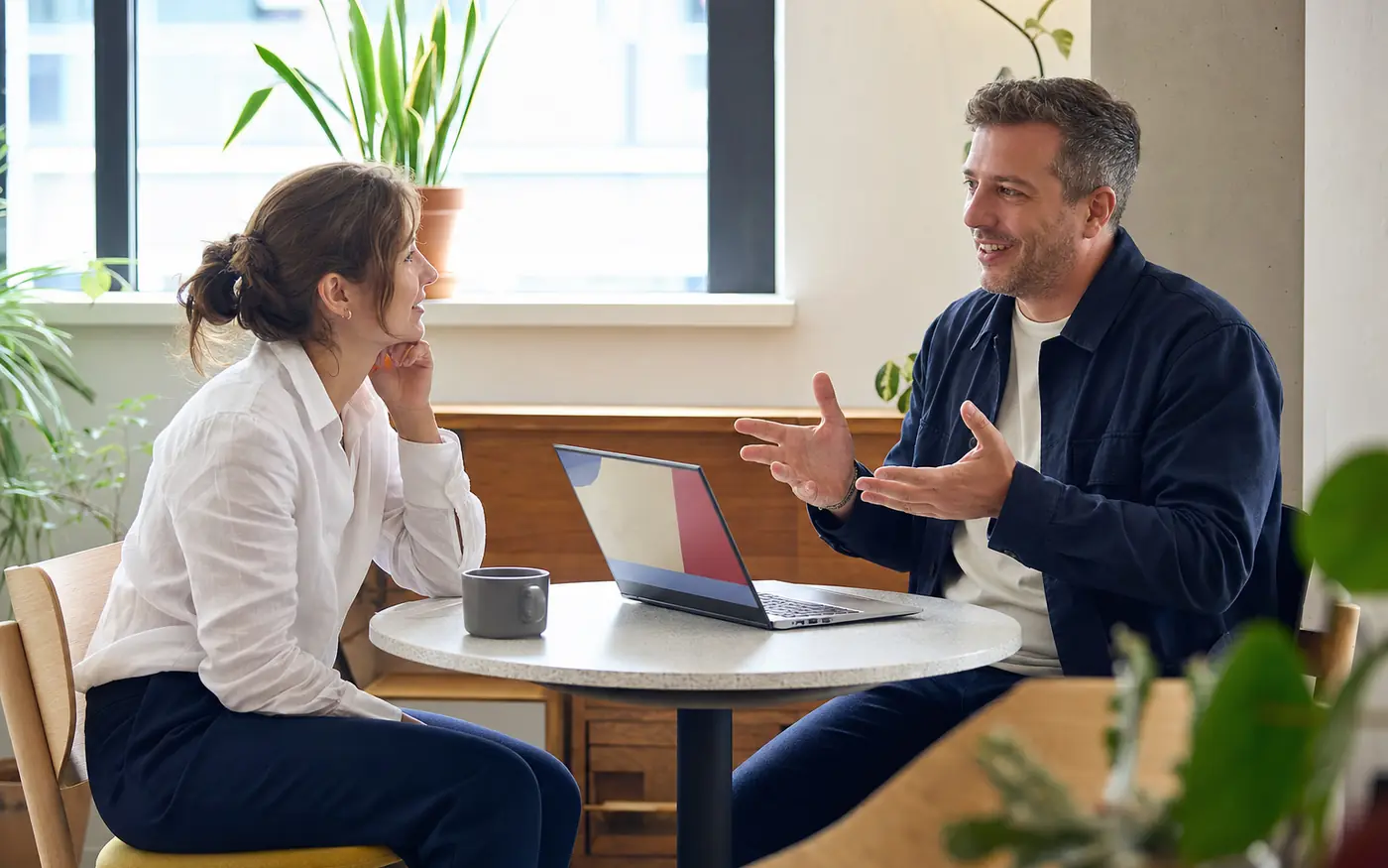 Two colleagues in a relaxed one-to-one conversation about their working styles, a laptop with a colourful profile between them